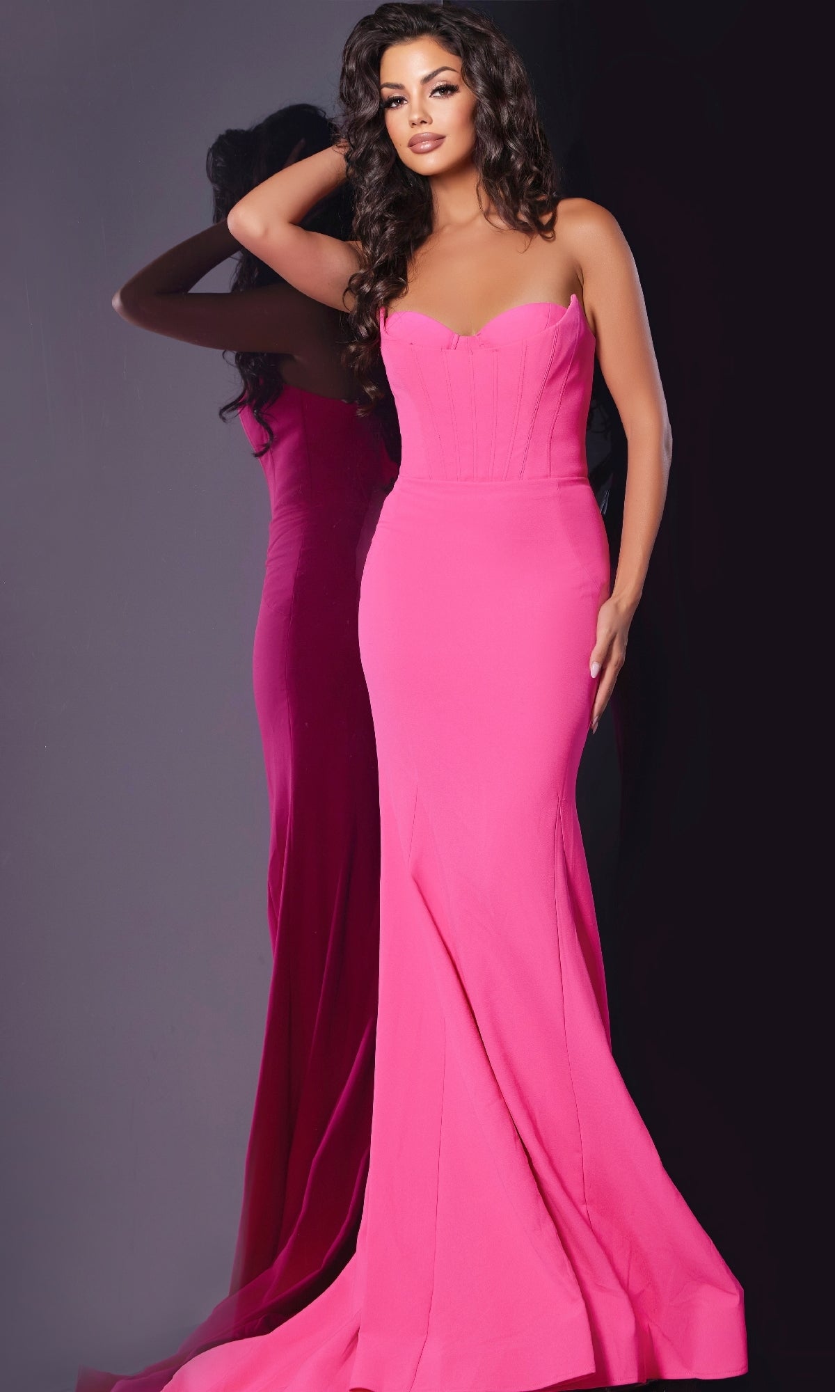 A woman in the strapless, fitted, floor-length pink Long Prom Dress: Jovani 38974 stands by a mirror, her left hand touching her hair and her right hand at her side.